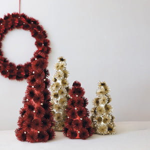 A set of decorative Christmas tree ornaments and a matching wreath made of layered paper flowers in deep red, champagne gold, and cream, displayed on a white surface against a light gray wall.