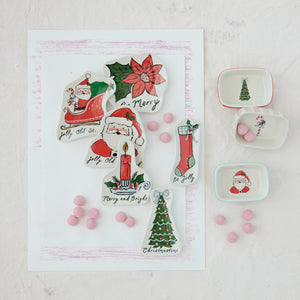A flat-lay of Christmas-themed ceramic tiles and small dishware on a white background. The set includes a large tile shaped like a Christmas tree with Santa, a poinsettia, a candle, stockings, and a Christmas tree, alongside three small square dishes with red trim featuring Santa, a candy cane, and a Christmas tree.