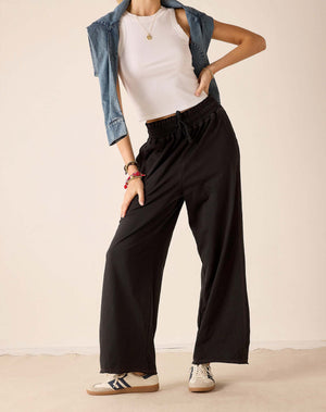 A model wearing black wide-leg drawstring pants, styled with a white tank top, an open blue denim jacket, and white sneakers, set against a plain beige studio background.