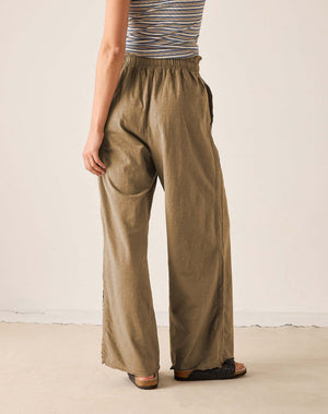 A person shown from the back wearing wide-leg nutmeg brown pants with an elastic waistband, paired with a striped top and black sandals, against a plain off-white wall.