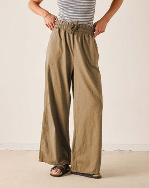 A person wearing loose, wide-leg olive khaki pants with an elastic drawstring waist, paired with a striped top and sandals, standing against a plain off-white wall.