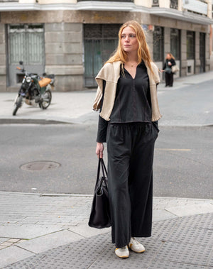 A woman stands on a city sidewalk wearing a black long-sleeve top and wide-leg black pants, with a beige cropped cardigan draped over her shoulders. She holds a black tote bag in an urban street setting.