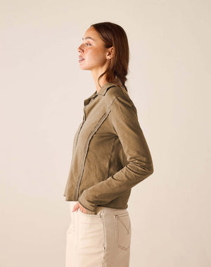 A woman shown in side profile wearing a cropped, v-neck cotton polo in a muted taupe-brown color, paired with light beige pants, against a plain light beige studio background.