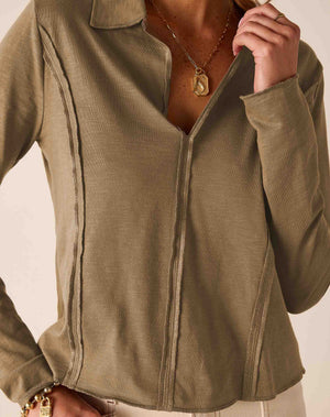 A close-up of a woman wearing a long-sleeve nutmeg brown V-neck shirt with a collar and vertical seam details, paired with a gold necklace and bracelet.
