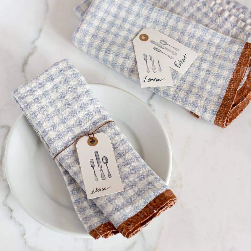 Blue and white gingham print handmade  linen napkins with brown trim on a marble surface with place setting details. Caravan collection. 