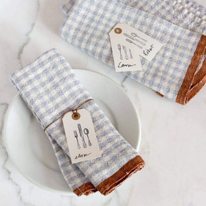 Blue and white gingham print handmade  linen napkins with brown trim on a marble surface with place setting details. Caravan collection. 