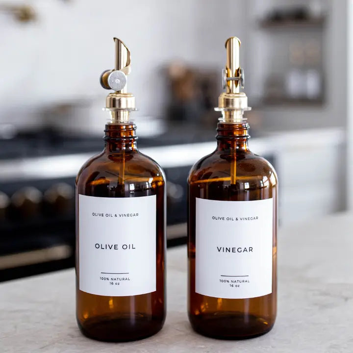 Two amber glass bottles with white labels, one labeled 'OLIVE OIL' and the other 'VINEGAR', each fitted with a gold-tone metal pour spout, set on a light countertop with a blurred kitchen background.