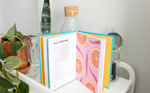 Open recipe book with colorful pages on a white surface with plants and jars.