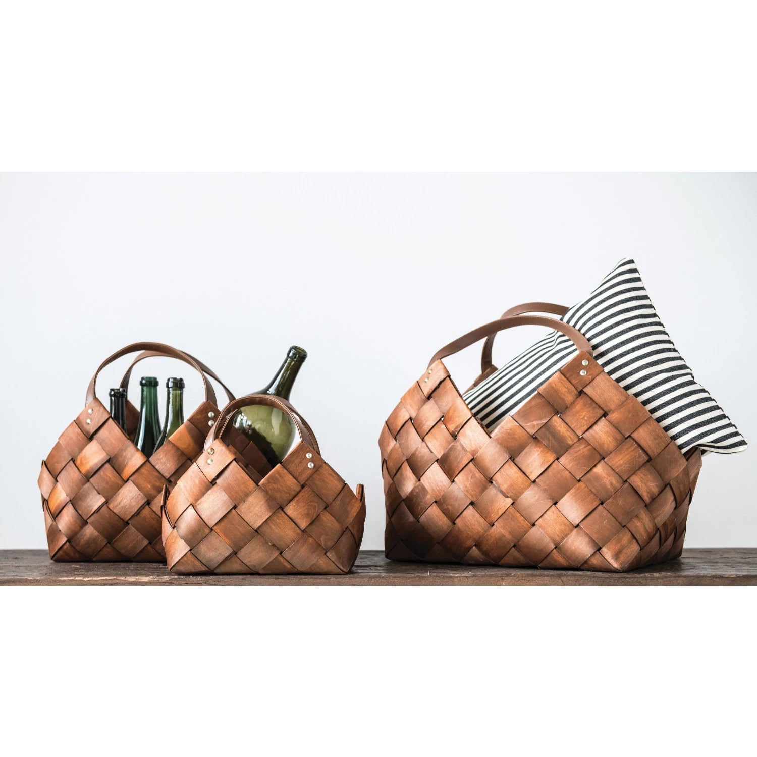Three filled woven brown seagrass baskets with leather handles on a wooden surface. 