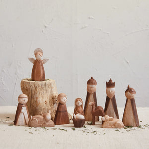 A set of wooden-like nativity figurines arranged on a light fabric surface with a textured off-white wall background. The set includes an angel standing on a small tree stump, baby Jesus in a manger, a donkey, and multiple wise men and shepherds in varying wood tones.