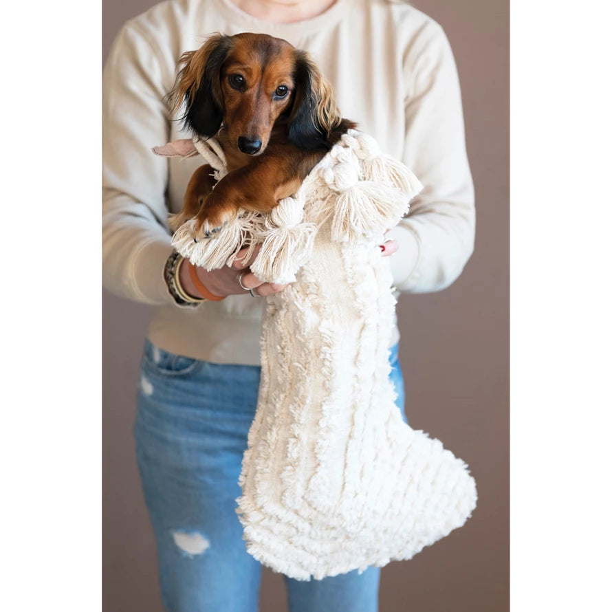 A person holding a white cotton tufted stocking with a dog inside, wearing a sweater and jeans.