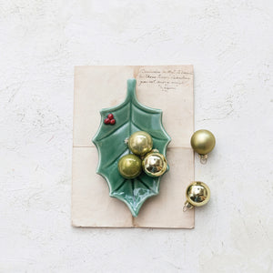 A glossy teal ceramic holly-shaped dish holding three gold Christmas ornaments, styled on top of a vintage handwritten card against a textured white background.