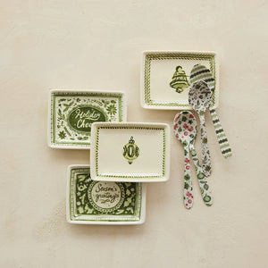 A set of four Christmas-themed ceramic dishes and three patterned spoons arranged on a textured beige background. The dishes are cream-colored with green borders and holiday designs including a Christmas tree, ornaments, and seasonal greetings, while the spoons feature green stripes and floral patterns.