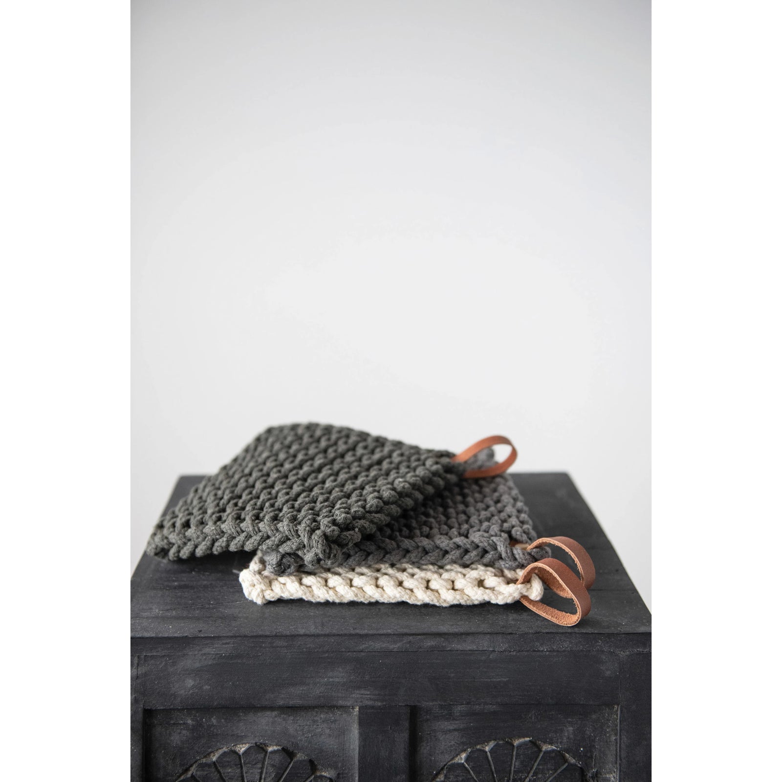 Modern crochet cotton potholders in neutral colors with leather loop strap; stacked on top pf black wood box. 